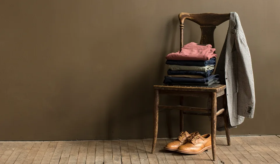 A stack of folded clothes on a wooden chair with a jacket over the back and a pair of tan shoes on the floor