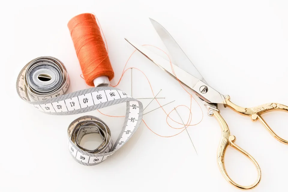 A soft tailor’s tape measure, a spool of orange thread and a pair of scissors on a white surface